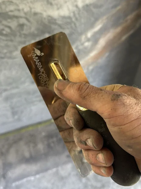 A plasterer's hand, grey with plaster dust, gripping a polished Marmorino trowel in front of a freshly plastered wall